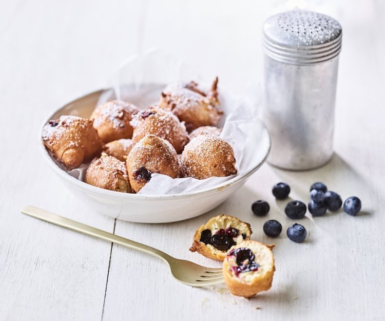 In een diep wit bord ligt een wit boterpapiertje met hierin enkele oliebollen. Er is eentje uitgeprikt en ligt op tafel doormidden gesneden, hier zie je duidelijk de blauwe bessen in zitten. Er is bloemsuiker over de oliebollen gestrooid voor een extra zoete toets. 