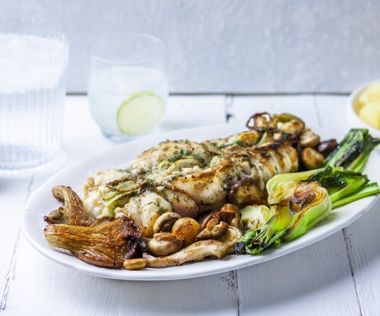 Ovengebakken zeeduivel ligt op een groot wit bord, geserveerd met gegrilde paksoi en champignons. Het gerechtje wordt geserveerd met gekookte aardappelen. Bovenaan staat ook ijskoud water met limoenschijfjes. 