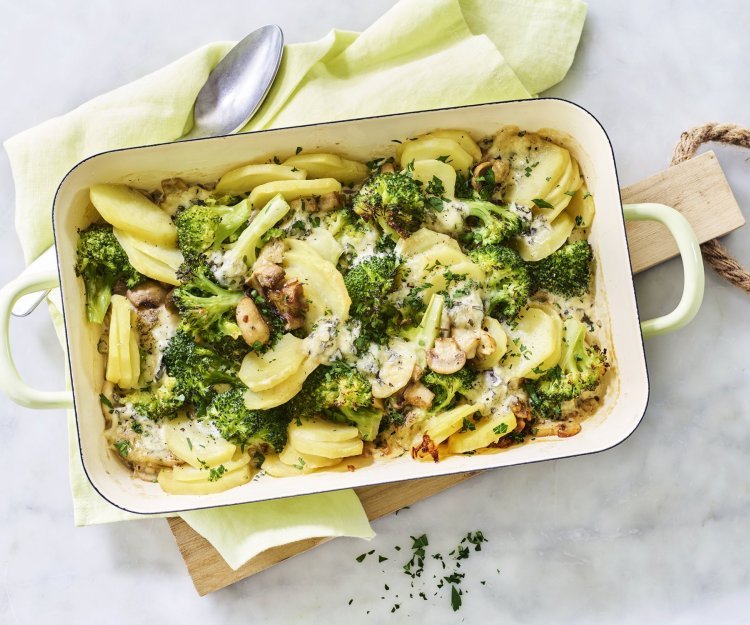 Een ovenschotel van aardappel, broccoli, champignons en blauwe kaas staat op het midden van het marmere aanrecht op een houten snijplank. Verse kruiden zorgen voor een extra groene touch!