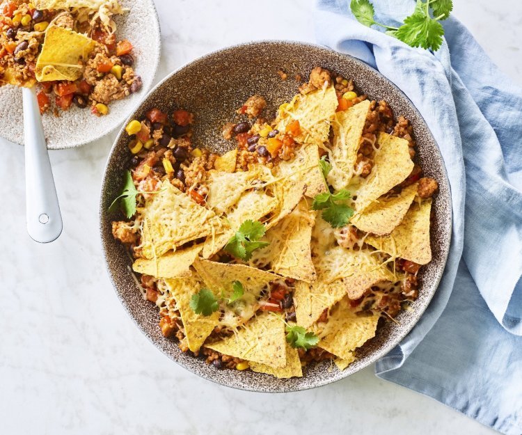 Onderaan de ronde ovenschaal ligt de chili con carne met bovenop de nachos. Er is al een portie uitgeschept op een wit bordje en staat langs de ovenschaal. Er ligt ook een blauwe servette en enkele blaadjes kroiander voor de kleur.