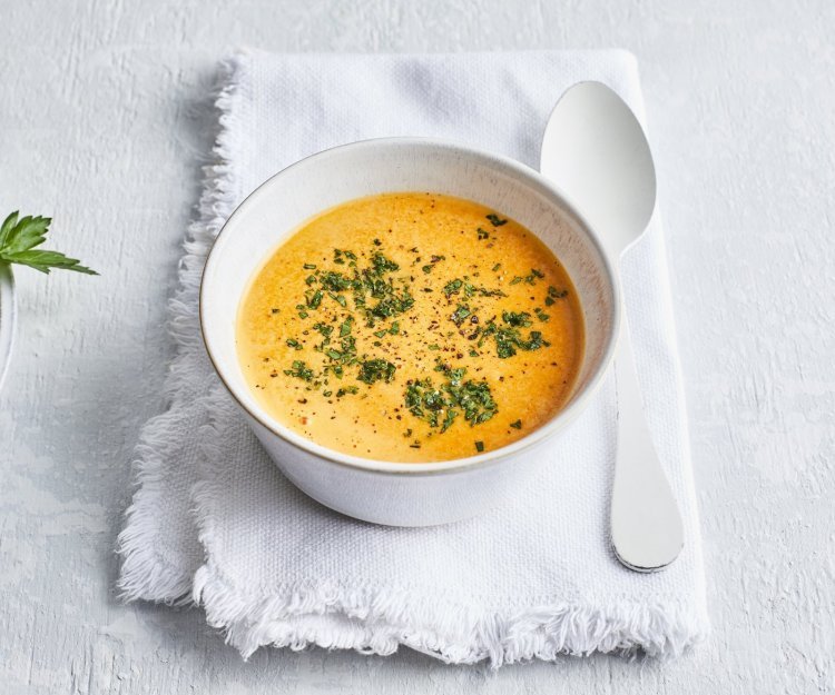 Deze paprikasaus zit een wit kommetje wat een mooi contrast geeft met de oranje kleur van de saus. Er ligt een witte lepel langs en de saus is afgewerkt met verse groene kruiden.