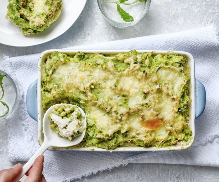 Eten is klaar! De ovenschotel staat op een witte dikke doek en een porseleine lepel schept de eerste portie uit de schotel. De parmentier heeft een mooie frisse groene kleur door de vele erwtjes. 