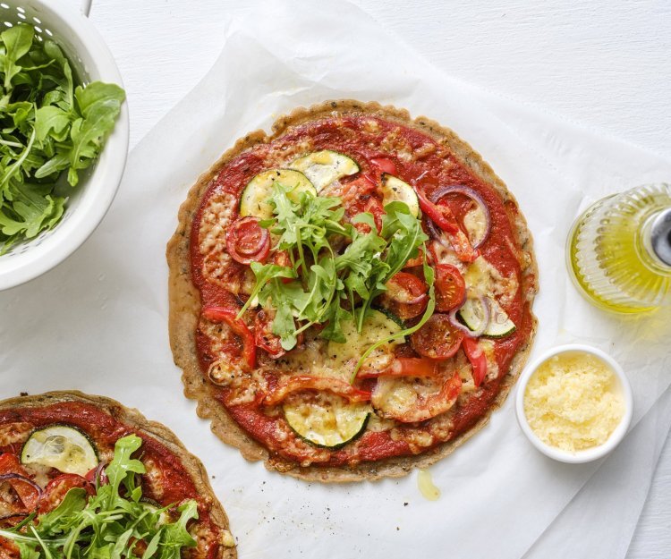 Op tafel staan twee klaargemaakte pizza's op een wit bakpapiertje. Hij is afgewerkt met verse rucola en wat olijfolie.