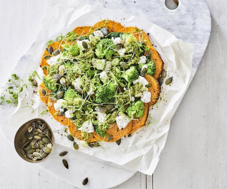 Klaargemaakt pizza van zoete aardappelschijfjes ligt op een snijplank van marmer. De pizza werd belegd met geitenkaas en groene groenten. 