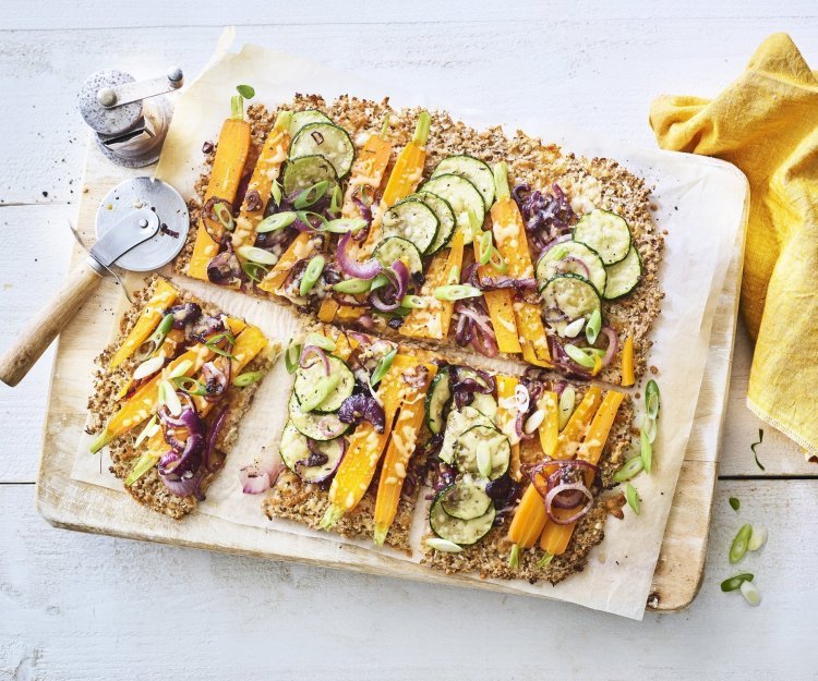 Een krokante bodem van bloemkool en walnoten waarop kleurrijke groentjes liggen zoals wortel, courgetteplakjes en rode ui. De plaattaart ligt op een houten snijplank waar een pizzasnijder langs ligt, de pepermolen en goudgele servette. 