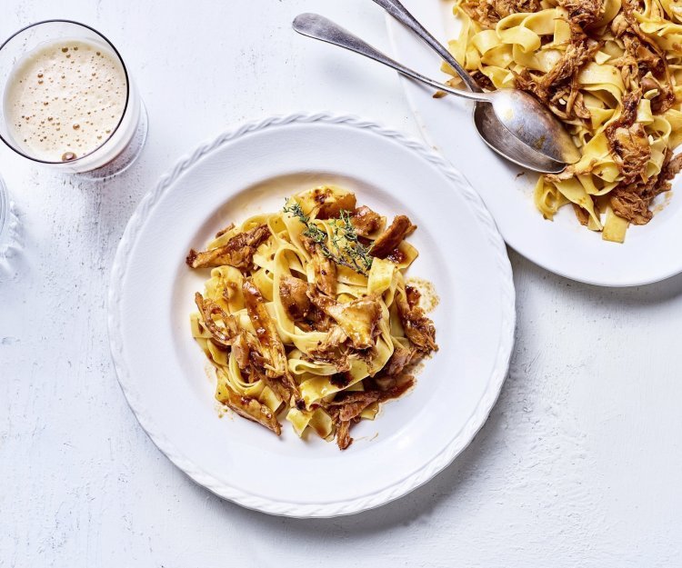 Op tafel staat een wit bord gevuld met tagliatelle en konijnenragout. Er staat een biertje langs, aangezien de ragout gemaakt is met Belgisch bier. 