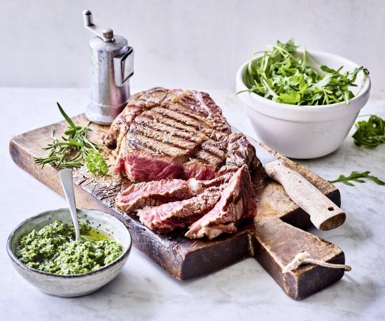 Een grote gegrilde steak ligt op een houten snijplank aangesneden. Er staat een potje homemade rucolapesto langs om meteen bij te scheppen bij je reepje vlees. Er staat ook een metalen pepermolen langs wie het graag wat pittiger wil.