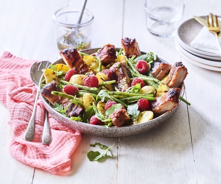 Een grote serveerschaal is gevuld met de aardappelsalade. Je ziet de framboosjes, boontjes en ribbetjes in de salade zitten. Er staat een torentje bordjes klaar met bestek om de salade te verdelen en op te eten. 