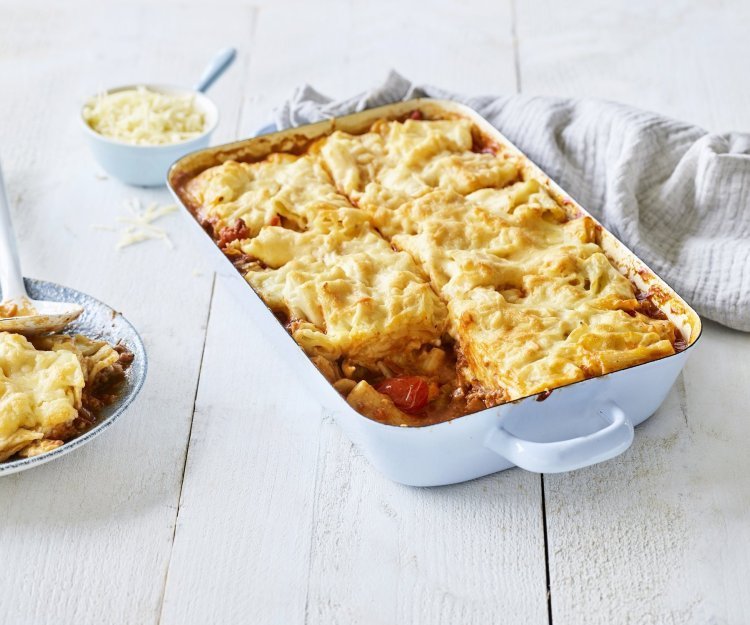 Een lichtblauwe ovenschotel staat op een houten witte tafel. De ovenschotel is gevuld met een mac & cheese én een tomatensaus, waardoor hij eerder weg heeft van een lasagne. Er is al 1 portie uitgeschept en wil al beginnen aan de volgende. Laat het niet koud worden. 