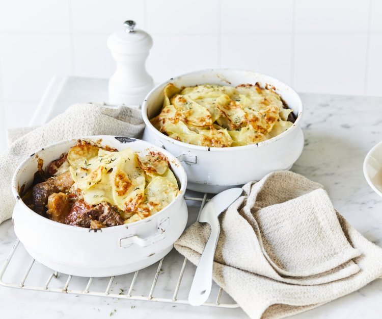 Twee witte stoofpotjes sttaan op een witte houten tafel en zijn gevuld met traag gegaard rundsvlees. De stoofpot is afgedekt met een laagje van aardappelschijfjes. De gratin is mooi goudgeel gebakken. Er staat een pepermolen langsheen de kookpotten, servette en grote lepel.