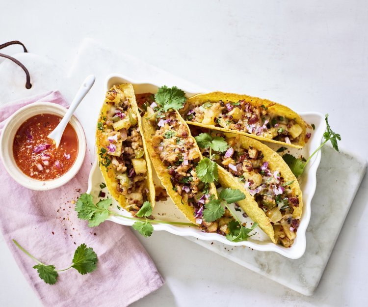 Vier tacos liggen klaar in een ovenschaal om aan te vallen. De pittige tomatendip staat links van de schotel op een roze servette. Het gerechtje is afgewerkt met verse korianderblaadjes.