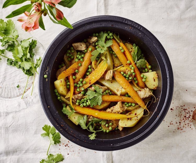 Een zwarte tajine waarin je kleurrijke ingrediënten vind. Denk maar aan wortel, aardappel, erwtjes en vlees. Er liggen nog wat verse kruiden naast om en een bloemetje voor de juiste sfeer in het beeld te krijgen. 