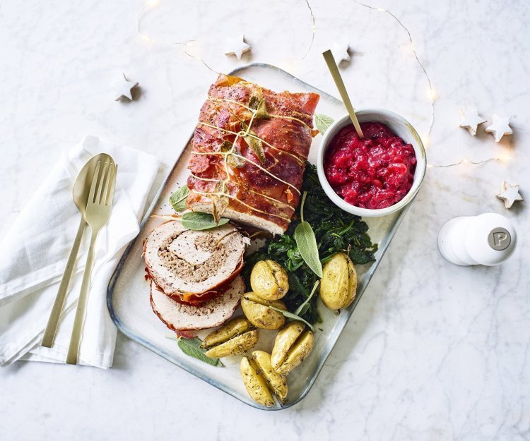 Een mooi aangesneden varkensrollade ligt op een rechthoekig dienblad. De aardappeltjes werden net niet doormidden gesneden alvorens ze de oven in gingen met een kruidentakje. De veenbessencompote staat langs de rollade die zorgt voor een intense kleur in deze feestmaaltijd. Goud bestek ligt langs het gerecht. 