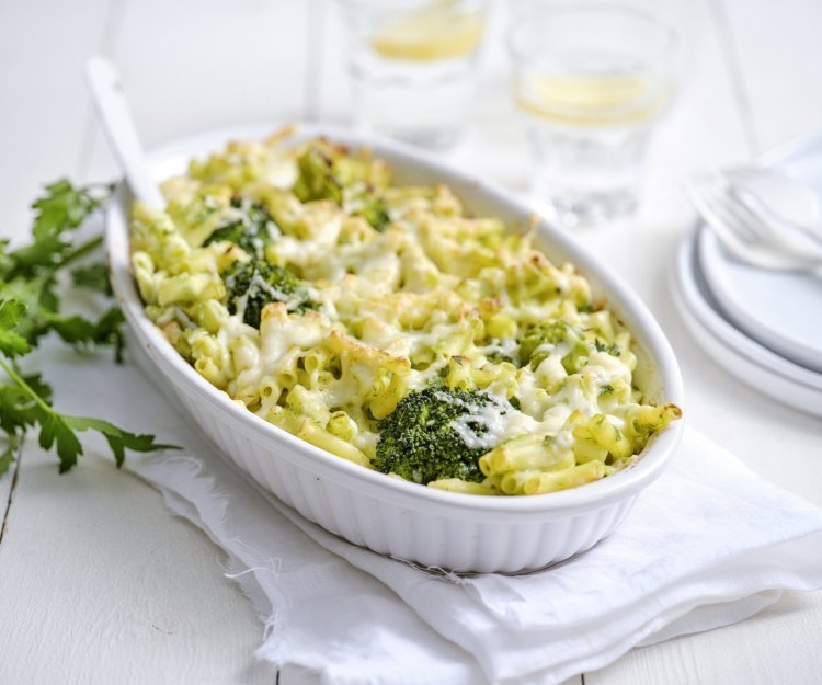 Een grote ovenschaal staat op een houte witte tafel. Hij staat op een wit lapje stof. De ovenschotel is gevuld met macaroni, broccolie en witte bloemkoolsaus. Er staan enkele bordjes langs met wit bestek om direct aan te vallen. 