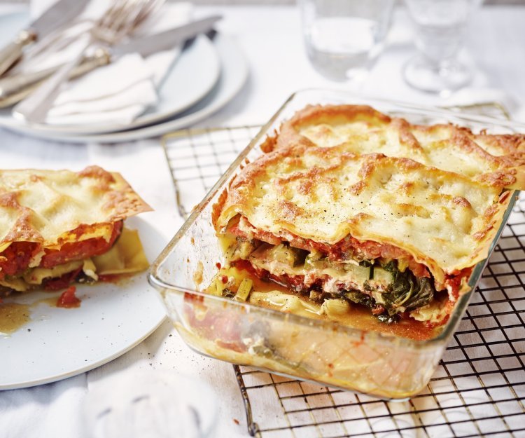 De lasagne staat op tafel in een glazen doorzichtige ovenschotel. Hij is aangesneden zodat je mooi de laagjes ziet van pasta, groenten en saus. De portie die is uitgeschept ligt op een bordje er naast.