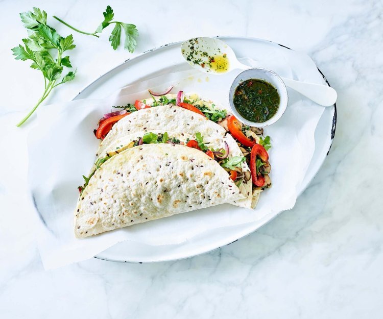 Vegetarische quesadilla gevuld met geroosterde knolselder, champignons, paprika en Brugge Oud. 2 stuks liggen gepresenteerd op een ovaal wit bord, samen met de vinaigrette in een potje apart. Takje verse koriander ligt er langs voor extra groen en sfeer aan het beeld mee te geven.