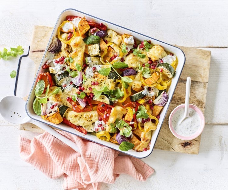 Op een witte tafel staat een houten snijplank met hierop een vierkante ovenschotel. Hij is gevuld met zoete aardappel, courgette, paprika en rode ui. Er is ook Berloumi gebruikt in dit gerechtje. 