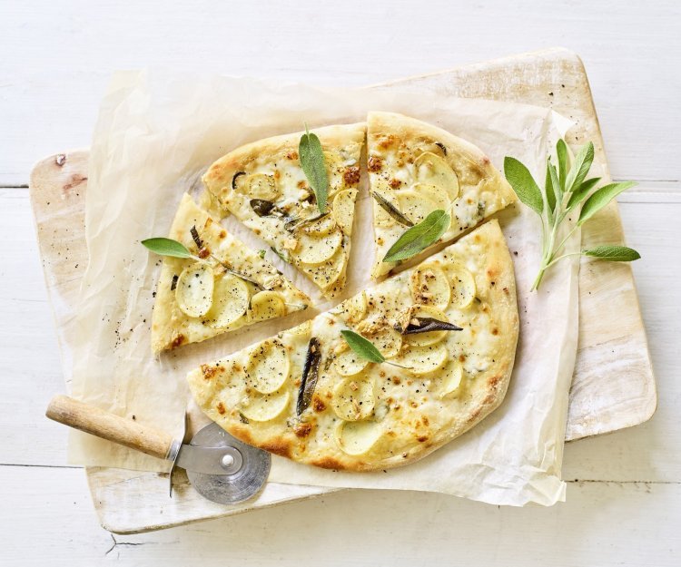 Een pizza belegt met mozzarella, plakjes aardappel en salie ligt op een wit boterpapiertje. Er ligt een pizzasnijder langs de pizza en nog enkele verse blaadjes salie. 