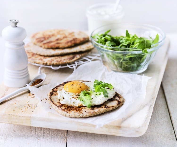 Het yoghurtbrood ligt op een houten snijplank. Hierop liggen meerdere ingrediënten om aan de slag mee te gaan, denk maar aan rucola, peper, extra yoghurt. 