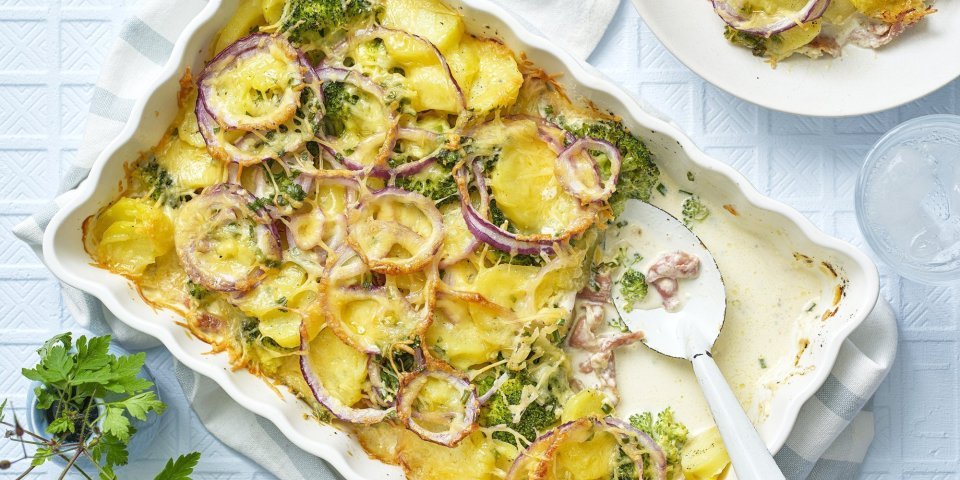 Een ovenschotel staat op tafel, waaruit al 1 hoek is uitgeschept op een bord. De ovenschotel bestaat uit krokante aardappelschijfjes, zoals een aardappelgratin, maar er is ook broccoli, rode ui en gerookte ham aan toegevoegd. Er staat een vaasje langs met een bloemetje en het tafelkleed is wit met ruitjes. 