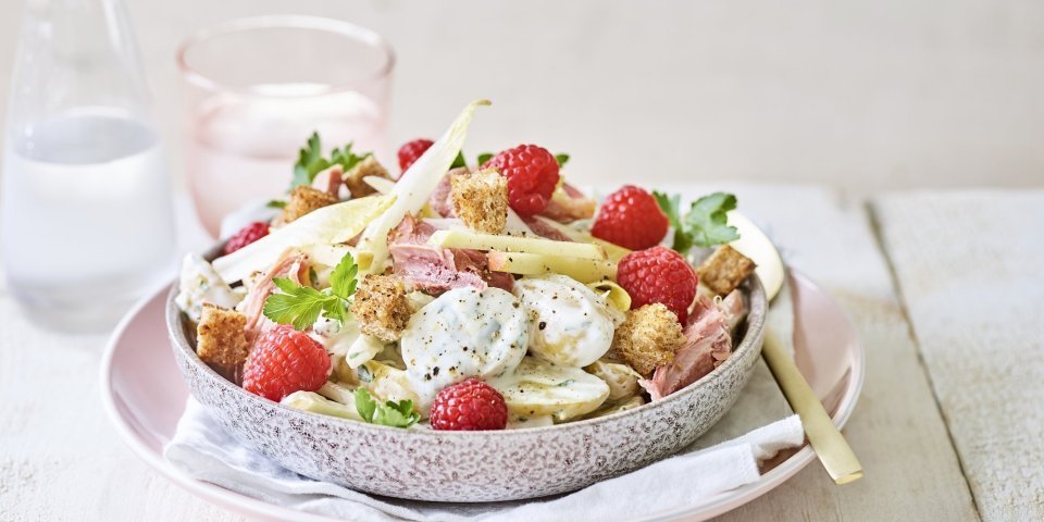 Een rijkelijke aardappelsalade is geserveerd in een grote kom. Je ziet de aardappel met de dressing, stukjes Breydelham, framboosjes en appel. De kom staat op een pastelkleurig roos bord. Er staat ook een karaf water langs met een glas in dezelfde tint als het roze bord. 