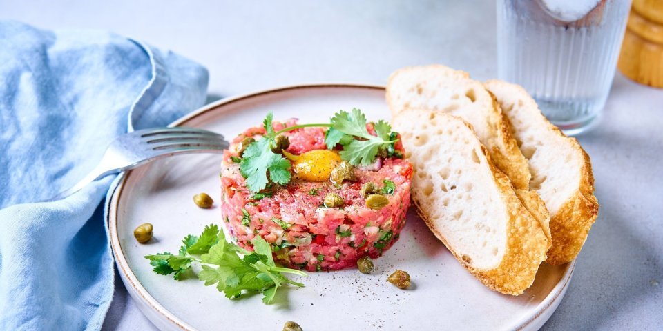 Een verse steak tartaar ligt in een mooi rondje op een bord. Bovenop ligt een kwarteleitje te blinken. 