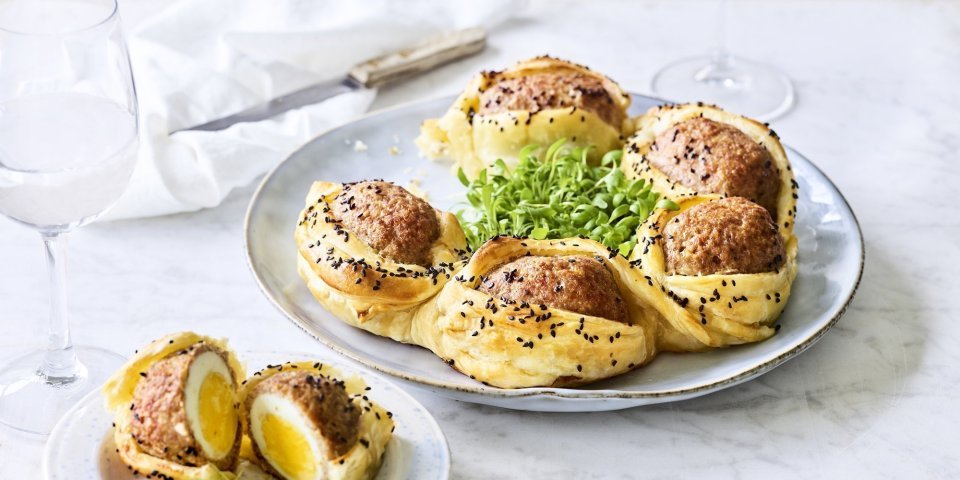 Op tafel staat een bord waarop een bladerdeegkrans ligt. Er zitten grote gehaktballen tussen gevluchten met hierin een gekookt eitje. Het bladerdeeg is afgewerkt met zwarte sesamzaadjes wat zorgt voor een leuke spikkeling.