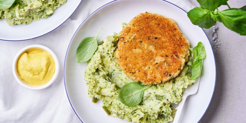 Op tafel staan 2 witte borden met een blauw randje. Hierin ligt een schep broccolipuree met een kaasburger en enkele blaadjes basilicum. Er staat ook een potje mosterd op tafel. 