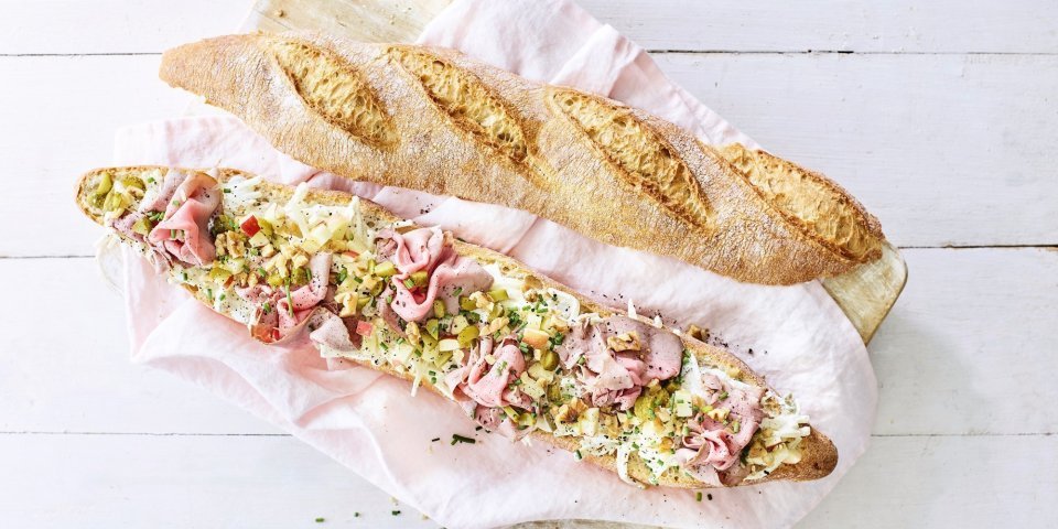 Een groot stokbrood ligt in de lengte opengesneden op een houten plank. Het broodje is belegd met een slaatje van knolselder, plukjes rosbief, stukjes appel en walnoten.