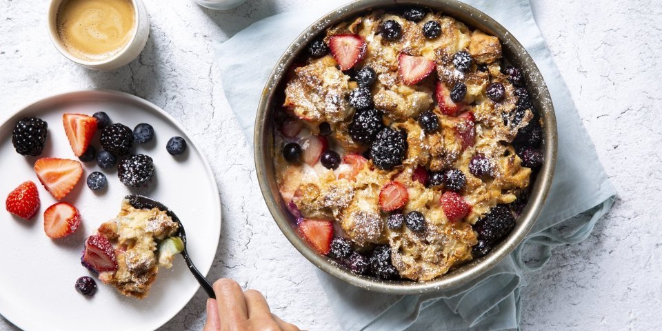 Homemade broodpudding met rabarber en zomerbessen staat op tafel. Een vrouwelijke gast schept haar een portie uit om volop te genieten van dit dessert met een kopje koffie. 