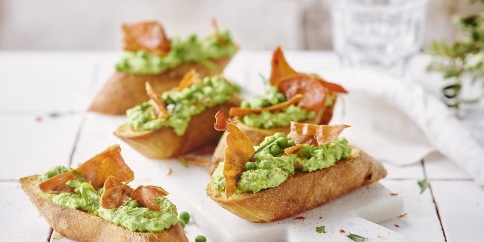 Vijf klaargemaakte bruschetta liggen op tafel. De spread van geitenkaas en erwten kleurt mooi groen, de bruchetta werd afgewerkt met een stukje krokant gebakken gedroogde ham. 