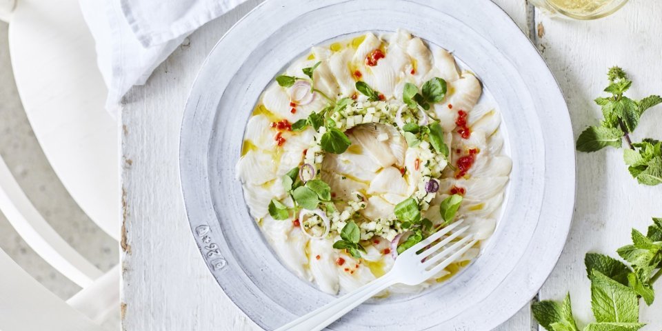 Ceviche van wijting met kleine stukjes koolrabi en appel er bovenop. Hij is geserveerd op een wit bord waardoor de groentjes nog extra opvallen. Er ligt verse munt langs en een witte servette. 