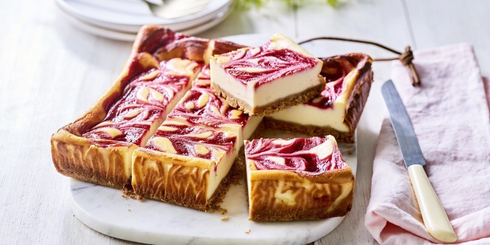 Een gebakken kaastaart staat op een marmere snijplank, het is wel een speciale cheesecake. Nl, de bovenzijde bevat frambozencoulis, waardoor er bovenaan een marmerpatroon is van de kaas en framboosjes. Echt een pareltje om naar te kijken. De taart is al aangesneden om onmiddelijk aan te vallen. 