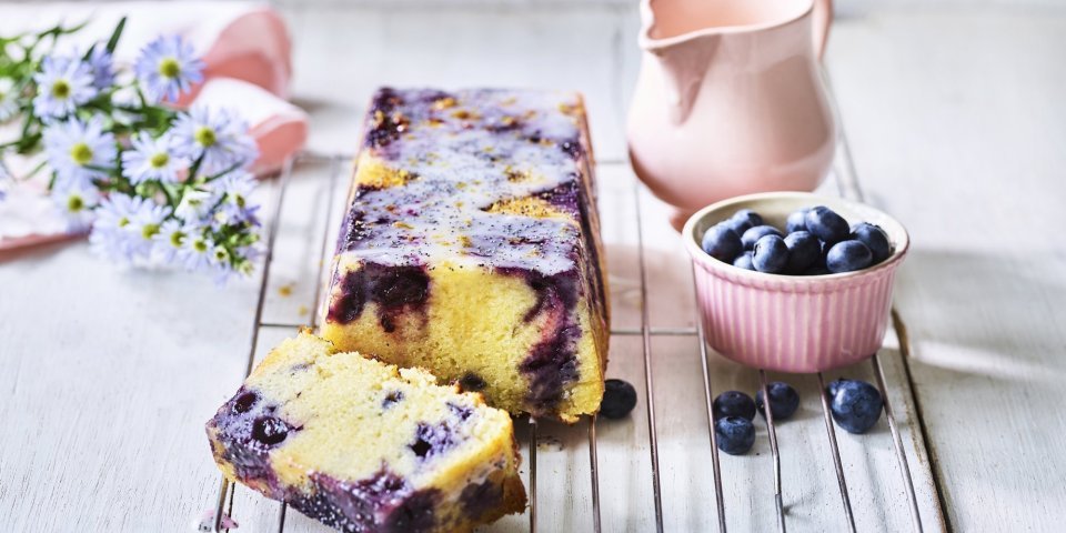 Op een bakrooster ligt de citroencake af te koelen, hij heeft een prachtige kleur door de blauwe bessen die tinten van paars achterlieten in de smeuige cake. Er staat nog een extra portie blauwe bessen naast voor extra fruitplezier.