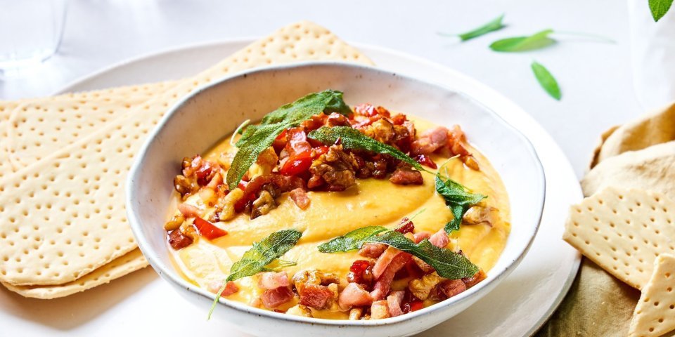 Er staat een diep bord op tafel, waarin de gemixte butternut met geitenkaas ligt. De dip is afgewerkt met stukjes gebakken spek, salie, noten en kruiden. Hij is prachtig oranje. Wij serveren hem met lange crackers. 