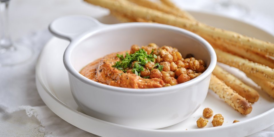 Een wit porseleine potje is gevuld met een oranjekleurige tapenade. Het is er eentje van zoete puntpaprika met hierop geroosterde kikkererwten. Er liggen broodstengels langs om te dippen. 