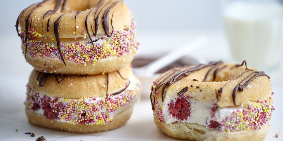 Drie donuts liggen op een witte stenen snijplank. De donuts zijn doormidden gesneden en gevuld met yoghurtijs met rode framboosjes, gerold door kleurrijke sprinkles en afgewerkt met gesmolten chocolade. In de achtergrond staat nog een glaasje melk.