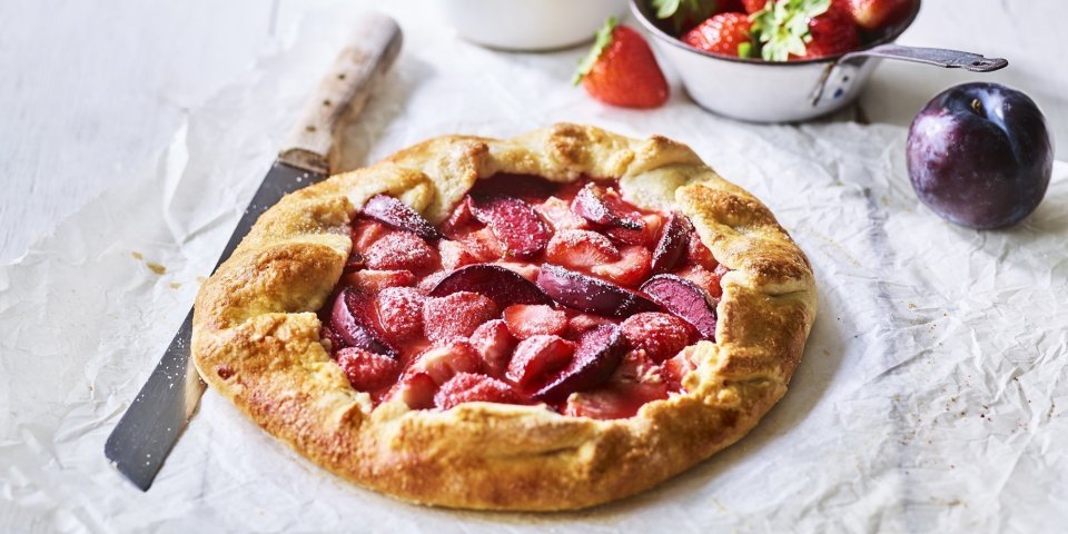 Een klaargemaakte galette staat op een houten witte tafel. Hij is prachtig goudgeel gebakken en het fruit oogt zoet-rode tinten. Er liggen nog wat aardbeien langs, alsook een paarse pruim.