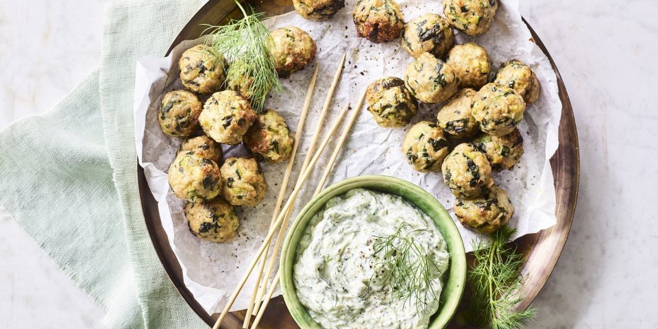 Op het aanrecht staat een houten ronde serveerschaal met een wit boterpapiertje op. Hierop liggen kleine gehaktballetjes met groene stukjes in van de courgette en spinazie. De dip op basis van tzatziki staat ook op de schaal. Het gerecht is afgewerkt met enkele takjes dille. 