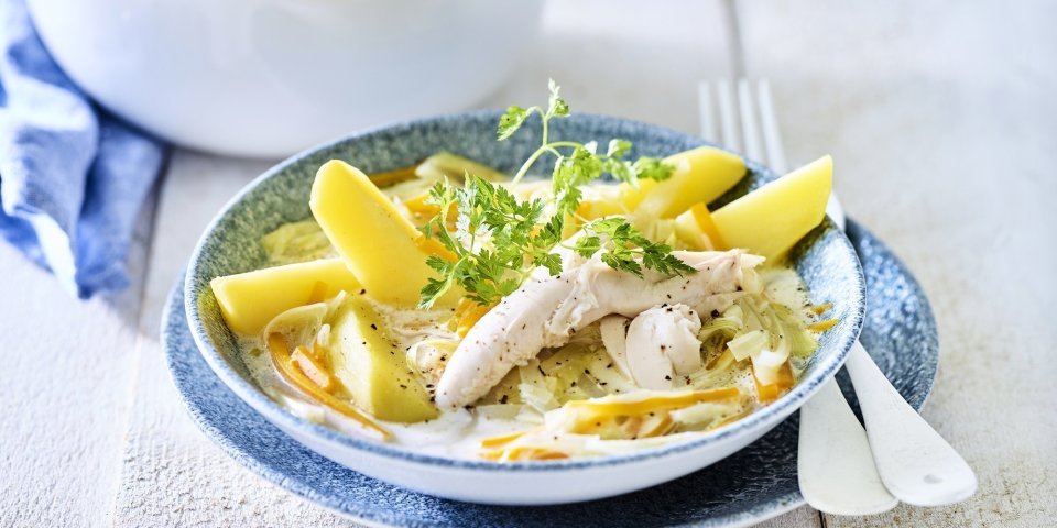 Op een witte houten tafel staat de kookpot vol waterzooi, er is een bordje uitgeschept met hierop de gekookte aardappelpartjes geschikt. Het gerechtje is afgewerkt met een takje verse kervel. 