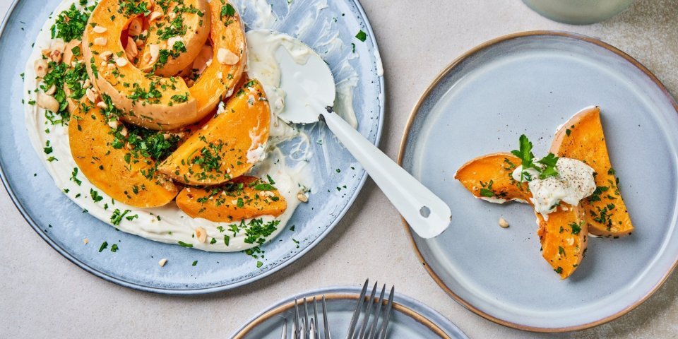 Er staat een lichtblauw-grijs bord waarop een kaasspread is uitgesmeerd. Hierop liggen stukken geroosterde butternut. Het recept is afgewerkt met fijngehakte peterselie en noten.