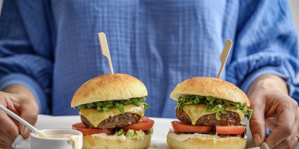 Een vrouw staat in beeld met haar romp draagt een plateau met hierop twee hamburgers. De hamburgerbroodjes hebben een mooi goudgele kleur, ze werden gemaakt met aardappelpuree in het deeg. Verder zijn de burger belgd met een schijfje vlees, kaas, tomaat, sla en saus natuurlijk. 