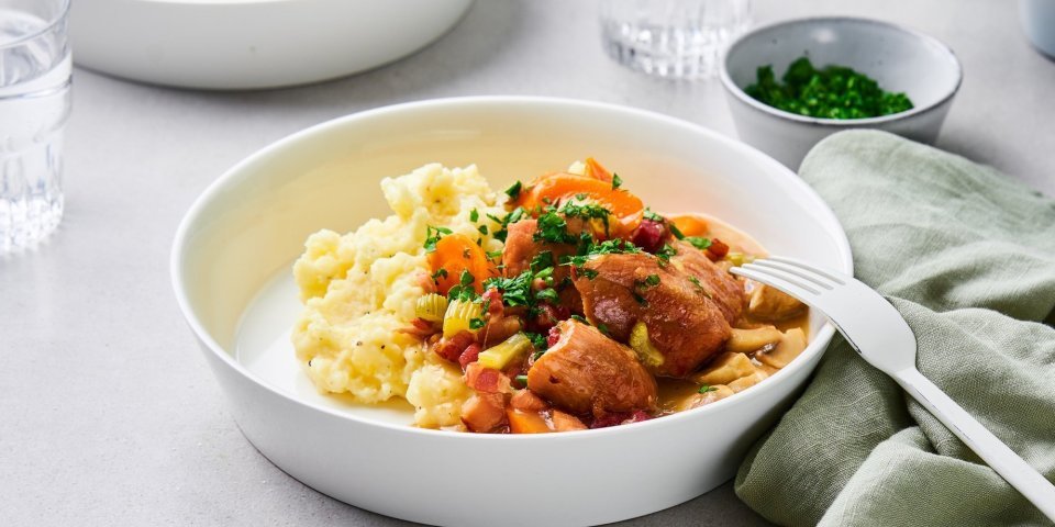 Op tafel staan twee uitgeschepte porties van het hoofdgerecht. Een schep heerlijke puree met kalfsfricassee. In de fricassee zitten spekblokjes, selder en wortelstukjes. Het gerechtje is afgewerkt met verse peterselie. Er ligt een licht groene servette langs en wit bestek.