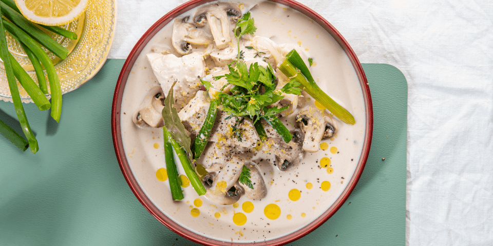 Kip gegaard in melk staat op tafel in een diep beige bord met bordeau randje. Er liggen lente-uitjes en champignons bij. Het bordje staat op een blauwgroene onderlegger. 