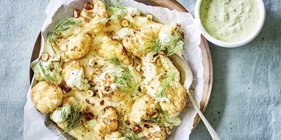 Puree van knolselder is uitgeschept op een groot lichtbruin bord. Hierop zijn de geroosterde bloemkoolroosjes geserveerd met wat drizzle van de groene kruidendressing, noten en verse dille. De overige kruidendressing is in een witte potje en staat langs het bruine bord, samen met nog wat gehakte noten in een ander potje. Alles staat op een een plaid in een blauwgrijze kleur.