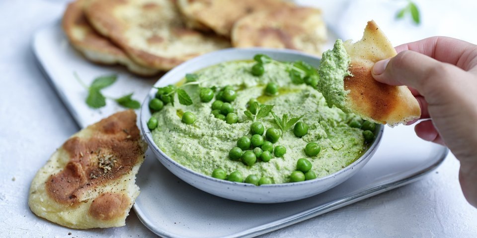 Krokant gebakken platbrood ligt op een serveerschaaltje waarbij een dip van erwten en roomkaas in geserveerd. De dip is gemixt en heeft een mooie groene kleur, om af te werken zijn er nog enkele verse erwtjes bovenop gestrooid. Een hand neem alvast een stukje platbrood om te dippen in gezonde groentedip. 