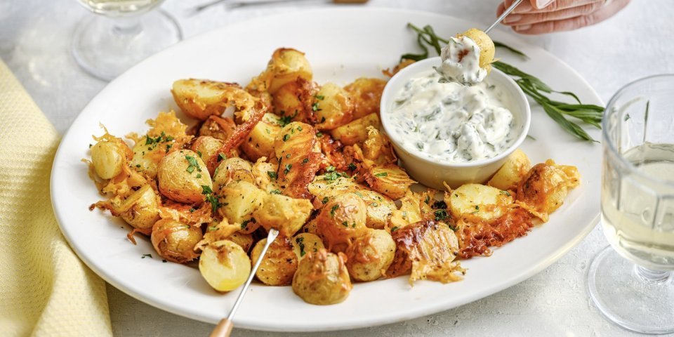 Een groot wit bord staat midden op de tafel, het is gevuld met heerlijke krokante aardappeltjes met kaas en een dipsausje van dragonmayonaise. Er is een hand die er eentje prikt en er staan twee glaasjes wijn. De aperitief is gestart. 