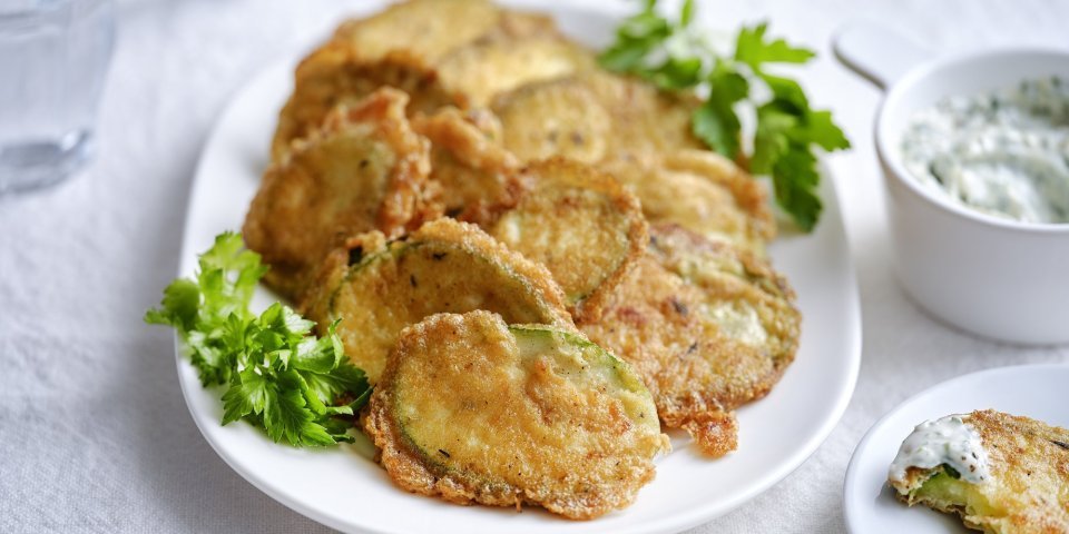 Krokante courgetteplakjes liggen op een bordje, afgewerkt met enkele blaadjes peterselie. De aioli staat langs het bordje.