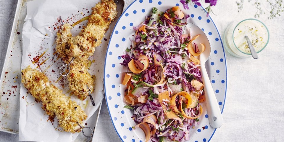 Krokante kipbrochette ligt nog de sidderen op een ovenplaatje. Daarnaast staat een kleurrijk groot bord met een salade op basis van rode kool, wortel en komkommer. De dressing op basis van yoghurt staat langs de salade.