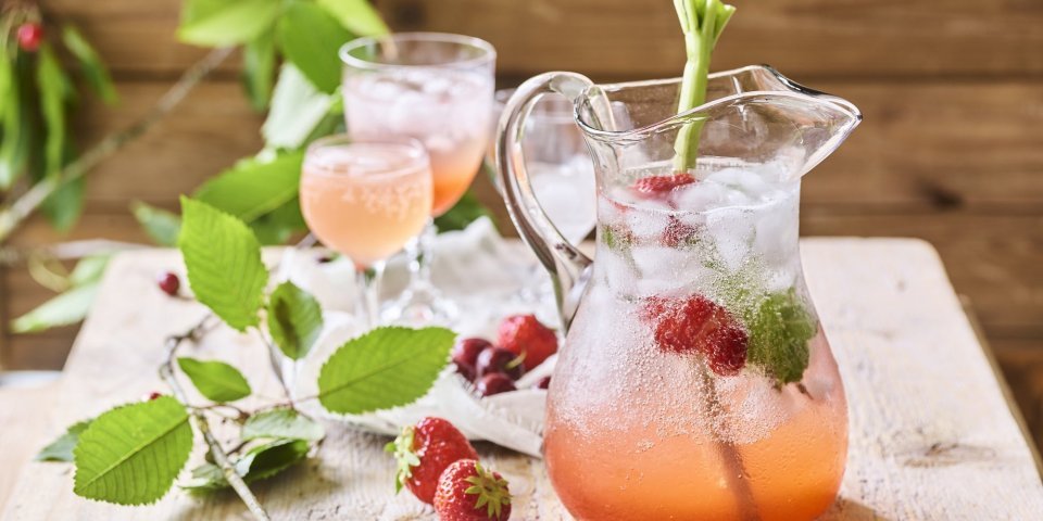 Er staat een karaf op een houten bruine buitentafel. Hij is gevuld met homemade limonade van rabarber en aardbeien. Er zitten ijsblokjes, muntblaadjes, aardbeien en een tak rabarber in de karaf. Er zijn twee glaasjes uitgeschonken. 